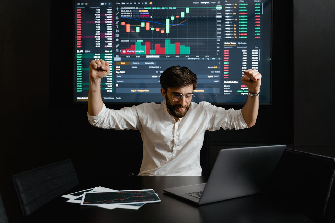 gallery-3 Businessman celebrates stock market success with hands raised in excitement at a trading desk.